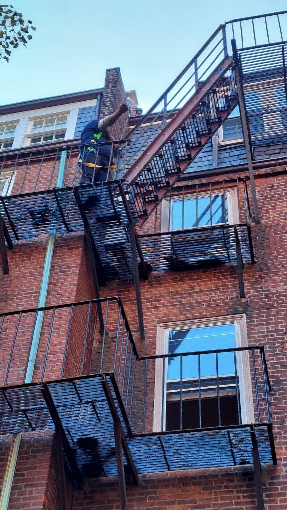 Technician in yellow safety harness performing elevated fire escape repair on a brick building