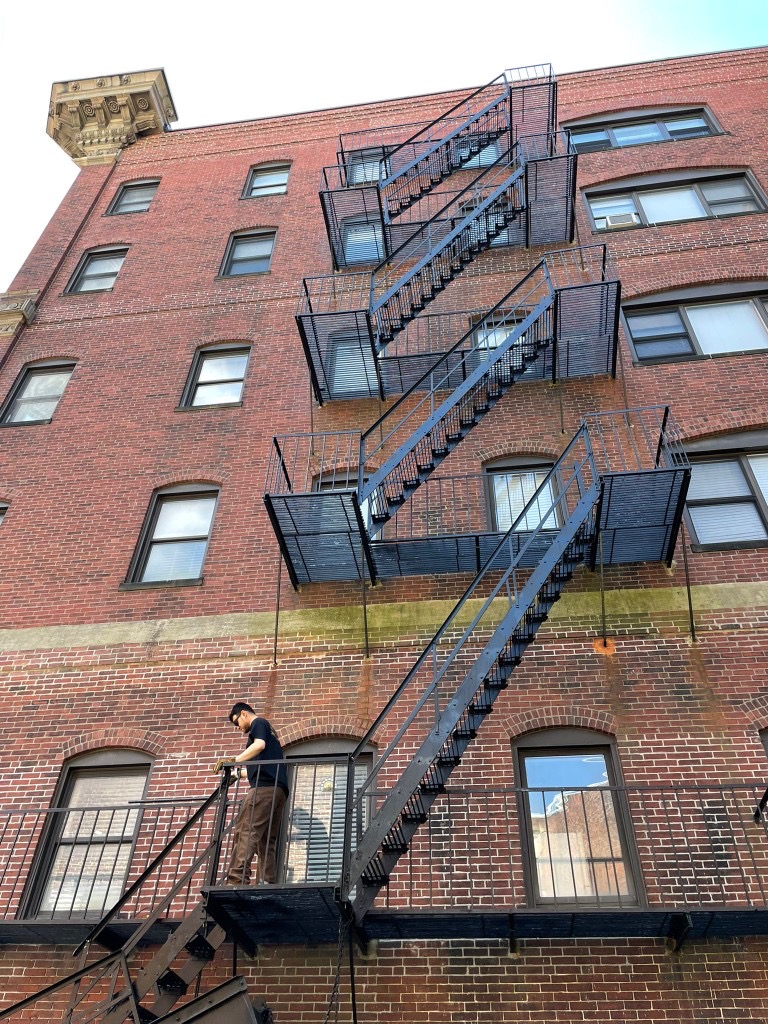 Technician inspecting a multi-story black fire escape on a red brick Boston building