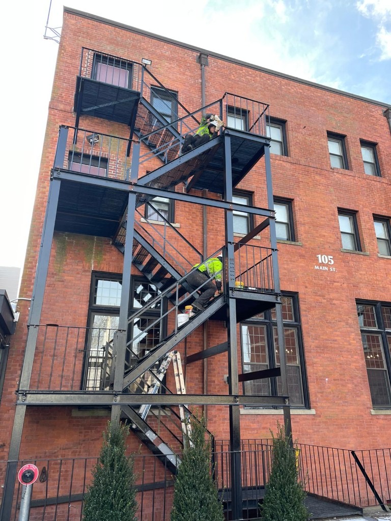 Two workers repairing a black fire escape on a Main Street brick building in Massachusetts
