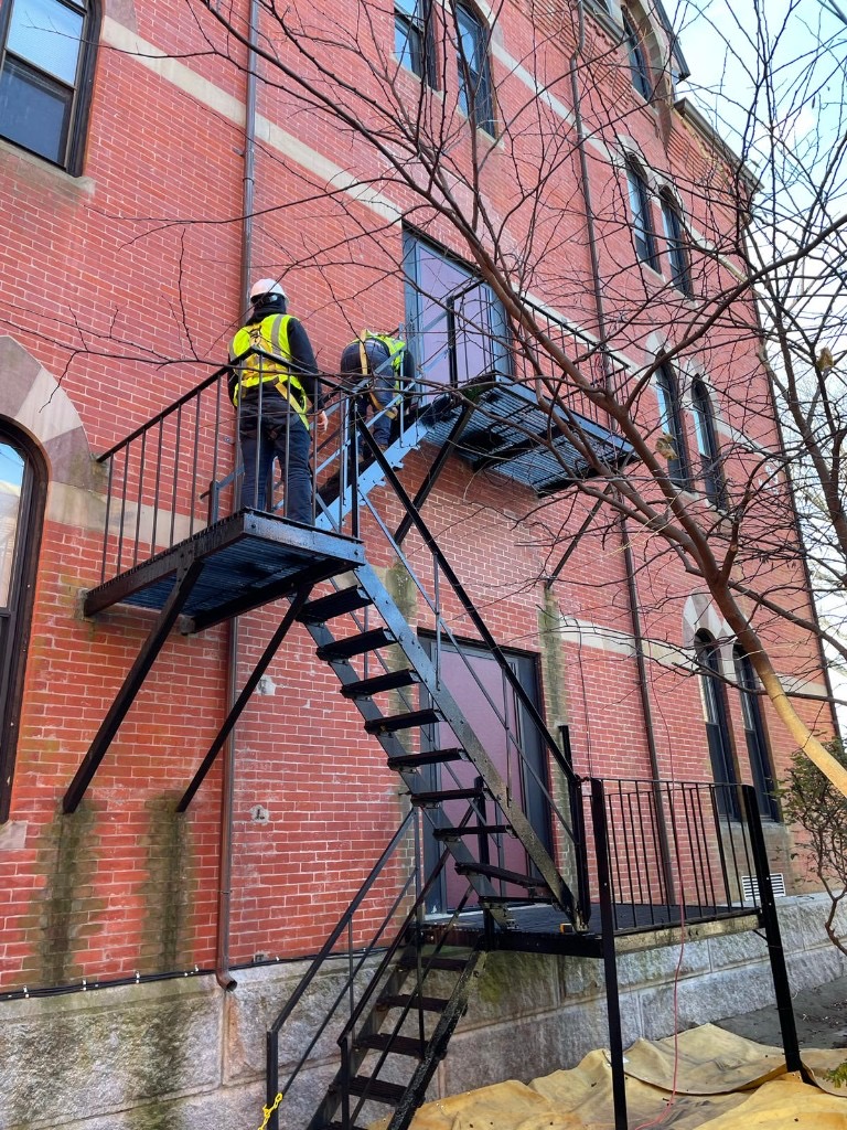 Boston Fire Escapes technicians in harnesses and hard hats working on a brick building fire escape