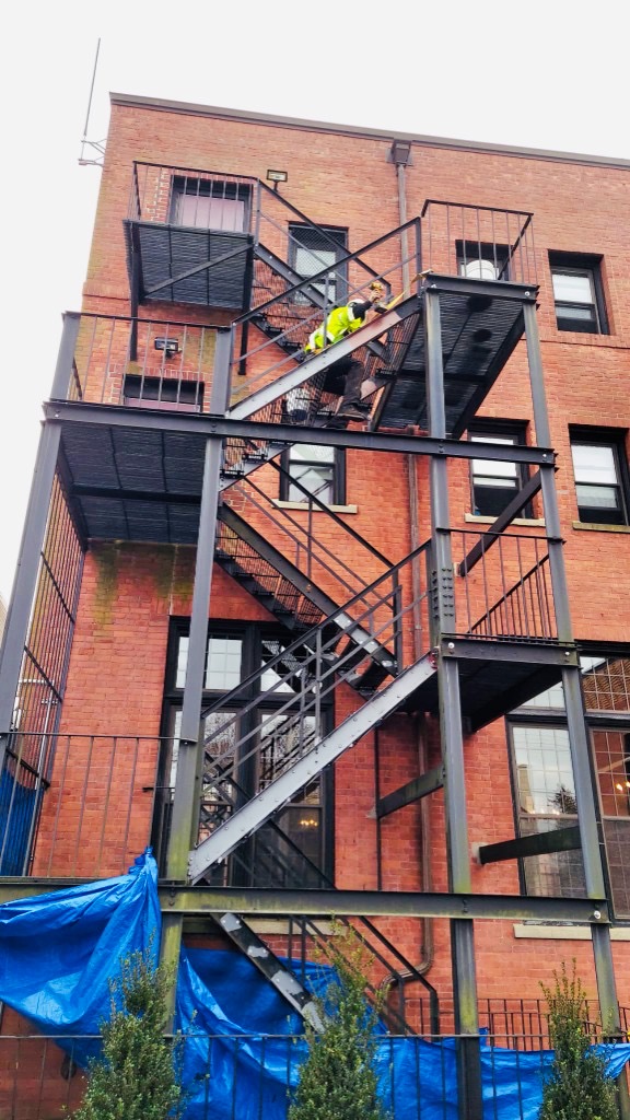 Technician performing structural fire escape repair mid-stair on an urban brick building