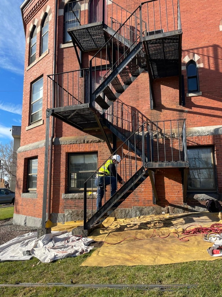Hard-hatted technician inspecting fire escape stair connections on a masonry building